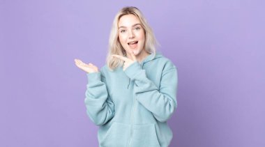 Güzel albino kadın neşeyle gülümsüyor ve yan taraftaki fotokopi alanını işaret ediyor, bir nesneyi gösteriyor veya reklamını yapıyor.