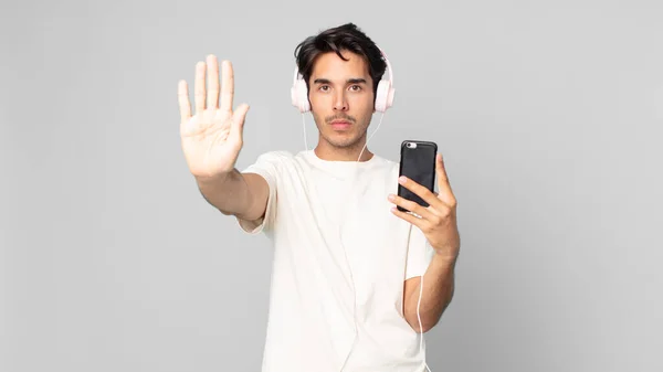 年轻的惊慌失措的男人看上去很严肃 带着耳机和智能手机展示着张开的手心 摆出了个停下来的姿势 — 图库照片