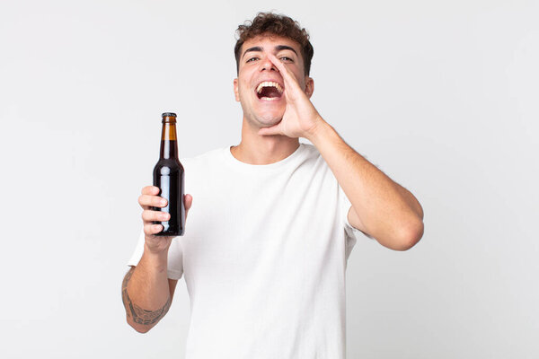 Молодой красивый мужчина чувствует себя счастливым, громко кричит, прижав руки к рту и держа пивную бутылку.