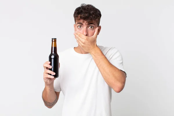 Młody Przystojny Mężczyzna Zakrywający Usta Dłońmi Szoku Trzymający Butelkę Piwa — Zdjęcie stockowe
