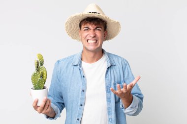 Genç ve yakışıklı bir adam sinirli, sinirli ve sinirli görünüyor. Elinde dekoratif kaktüs tutan çiftçi.