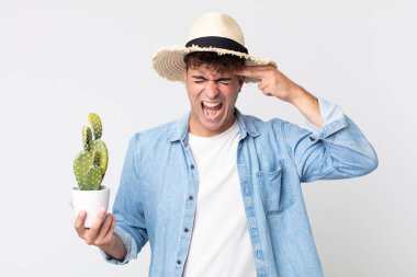Genç yakışıklı adam mutsuz ve stresli görünüyor, intihar jesti silah işareti yapıyor. Elinde dekoratif kaktüs tutan çiftçi.
