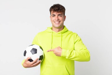 Genç yakışıklı adam neşeyle gülümsüyor, mutlu hissediyor ve bir tarafı işaret ediyor ve elinde bir futbol topu tutuyor.