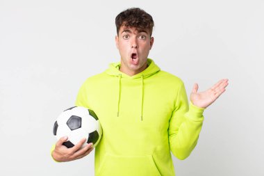 Genç yakışıklı adam şaşırmış ve şok olmuş görünüyor, çenesi düşmüş, elinde bir nesne, elinde bir futbol topu tutuyor.
