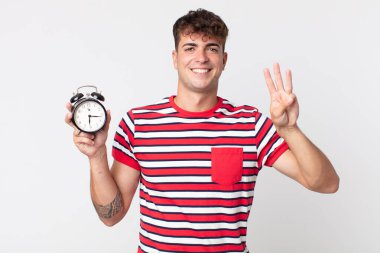 young handsome man smiling and looking friendly, showing number three and holding an alarm clock clipart