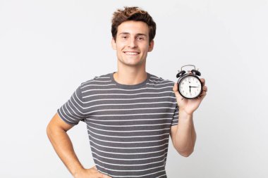 Genç yakışıklı adam mutlu bir şekilde gülümsüyor, eli kalçasında, kendinden emin ve elinde çalar saat tutuyor.