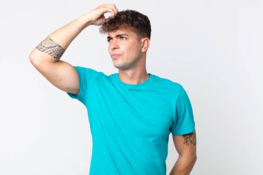 Genç yakışıklı adam şaşkın ve şaşkın hissediyor, başını kaşıyor ve yana bakıyor.