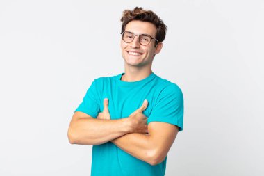 Genç yakışıklı adam kollarını kavuşturup mutlu bir şekilde gülüyor, rahat, pozitif ve tatmin edici bir pozla.