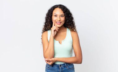 Güzel İspanyol kadın mutlu görünüyor ve gülümsüyor, elleri çenesinde, merak ediyor ya da soru soruyor, seçenekleri karşılaştırıyor.