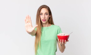 İnce, güzel bir kadın ciddi görünüyor. Avuç içini gösteriyor. El hareketi yapıyor ve elinde kahvaltı kasesi tutuyor.