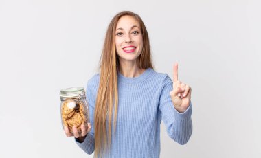 İnce, güzel bir kadın gülümsüyor ve arkadaş canlısı görünüyor. Bir numarayı gösteriyor ve elinde kurabiye tutuyor.