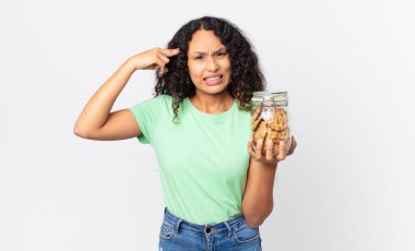 İspanyol güzel kadın şaşkın ve şaşkın hissediyor, deli olduğunu ve elinde kurabiye şişesi tuttuğunu gösteriyor.