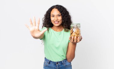 İspanyol güzel kadın gülümsüyor ve arkadaş canlısı görünüyor, 5 numarayı gösteriyor ve elinde bir kurabiye şişesi var.