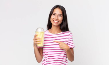 Genç İspanyol kadın neşeyle gülümsüyor, mutlu hissediyor ve kenarı işaret ediyor ve vanilyalı bir milkshake tutuyor.