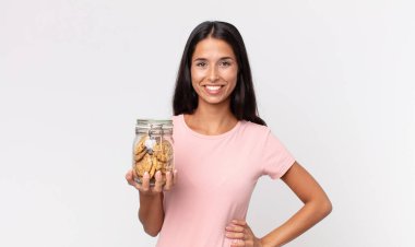 Genç İspanyol kadın mutlu bir şekilde gülümsüyor, eli kalçasında, kendinden emin ve elinde cam bir kurabiye şişesi tutuyor.