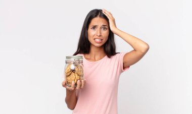 Genç İspanyol kadın kendini stresli, endişeli ya da korkmuş hissediyor. Elleri başının üstünde ve elinde cam bir kurabiye şişesi tutuyor.