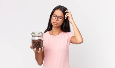 Genç İspanyol kadın şaşkın ve kafası karışmış hissediyor. Kafasını kaşıyor ve elinde kahve şişesi tutuyor.