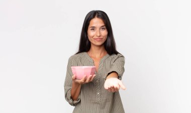 Genç İspanyol kadın mutlu bir şekilde gülümsüyor ve bir konsept sunuyor ve elinde boş bir kâse ya da demlik tutuyor.