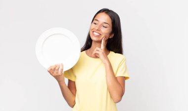 Genç İspanyol kadın mutlu bir şekilde gülümsüyor ve hayal kuruyor ya da şüphe ediyor ve elinde boş bir tabak tutuyor.