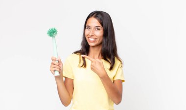 Genç İspanyol kadın neşeyle gülümsüyor, mutlu hissediyor ve bir yandan işaret ediyor ve elinde bir bulaşık fırçası tutuyor.