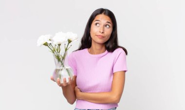 Genç İspanyol kadın omuz silkiyor, kafası karışık ve elinde dekoratif çiçekler tutuyor.