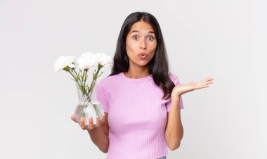 Genç İspanyol kadın şaşırmış ve şok olmuş görünüyor, çenesi düşmüş elinde dekoratif çiçekler tutan bir nesne tutuyor.