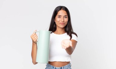 Genç İspanyol kadın mutlu bir şekilde gülümsüyor ve bir konsept sunuyor ve elinde kahve termosu tutuyor.