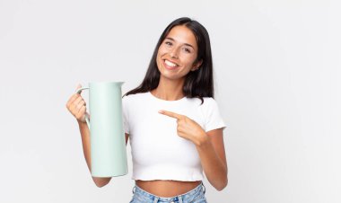 Genç İspanyol kadın neşeyle gülümsüyor, mutlu hissediyor ve bir tarafı işaret ediyor ve elinde kahve termosu tutuyor.