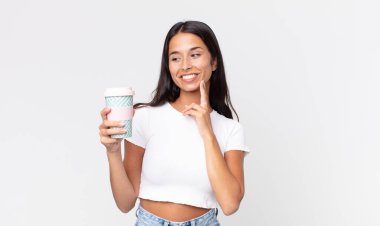 Genç İspanyol kadın mutlu bir şekilde gülümsüyor ve hayal kuruyor ya da şüphe ediyor ve elinde kahve kabını tutuyor.