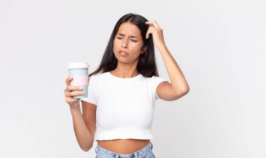 Genç İspanyol kadın şaşkın ve kafası karışmış hissediyor. Kafasını kaşıyor ve elinde kahve kabı tutuyor.