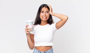 Genç İspanyol kadın stresli, endişeli ya da korkmuş hissediyor. Elleri başının üstünde ve elinde kahve kabı var.