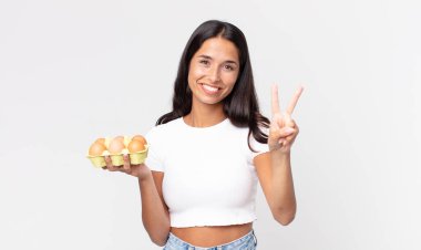 Genç İspanyol kadın gülümsüyor ve arkadaş canlısı görünüyor. İki numarayı gösteriyor ve elinde bir yumurta kutusu var.