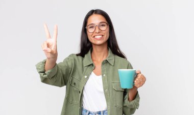 Genç İspanyol kadın gülümsüyor ve mutlu görünüyor, zafer ya da barış işareti yapıyor ve elinde kahve fincanı tutuyor.