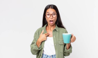 Genç İspanyol bir kadın ağzı açık, şok olmuş ve şaşırmış görünüyor. Kendini işaret ediyor ve elinde bir kahve fincanı tutuyor.