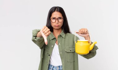 Genç İspanyol kadın kendini kötü hissediyor, baş parmağını kaldırıyor ve elinde bir çaydanlık tutuyor.