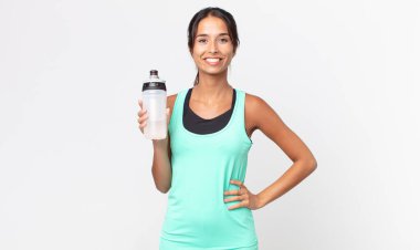 Genç İspanyol kadın mutlu bir şekilde gülümsüyor, eli kalçasında, kendinden emin ve elinde bir su şişesi tutuyor. fitness konsepti