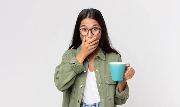 ショックを受けた若いヒスパニック系の女性が手で口を覆いコーヒーマグカップを持ち — ストック写真