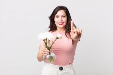 Güzel bir kadın gülümsüyor ve arkadaş canlısı görünüyor. Bir numarayı gösteriyor ve elinde dekoratif bir saksı tutuyor.