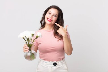 Kendine güvenen güzel bir kadın kendi gülüşünü işaret ediyor ve elinde dekoratif bir saksı tutuyor.