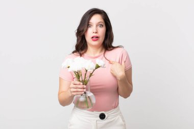 Güzel bir kadın şaşkın ve şaşkın görünüyor ağzı açık, kendini işaret ediyor ve elinde dekoratif bir saksı tutuyor.