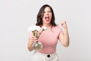 Güzel bir kadın sinirli bir ifadeyle bağırıyor ve elinde dekoratif bir saksı tutuyor.