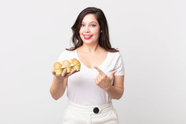 Güzel bir kadın heyecanlı ve şaşırmış görünüyor. Yan tarafı gösteriyor ve elinde bir yumurta kutusu tutuyor.