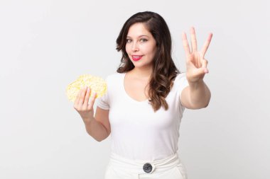 Güzel bir kadın gülümsüyor ve arkadaş canlısı görünüyor, üç numarayı gösteriyor ve elinde diyet pirinç keki tutuyor.