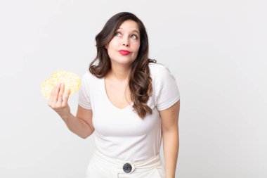 Güzel bir kadın omuz silkiyor, kafası karışık ve kararsız ve elinde diyet pirinç keki tutuyor.