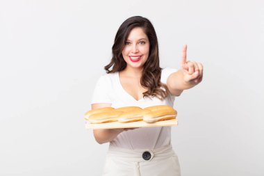 Güzel bir kadın gülümsüyor ve arkadaş canlısı görünüyor. Bir numarayı gösteriyor ve elinde bir ekmek tepsisi tutuyor.