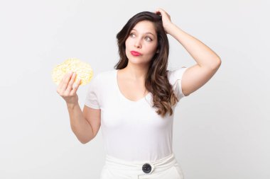 Güzel kadın şaşkın ve kafası karışmış hissediyor, kafasını kaşıyor ve elinde diyet pirinç keki tutuyor.