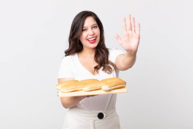 Güzel bir kadın gülümsüyor ve arkadaş canlısı görünüyor, beş numarayı gösteriyor ve elinde bir ekmek tepsisi tutuyor.