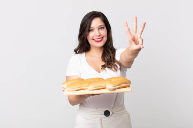Güzel bir kadın gülümsüyor ve arkadaş canlısı görünüyor, üç numarayı gösteriyor ve elinde bir ekmek tepsisi tutuyor.