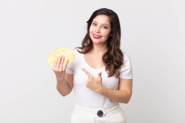 Güzel bir kadın neşeyle gülümsüyor, mutlu hissediyor ve bir tarafı işaret ediyor ve elinde diyet pirinç keki tutuyor.