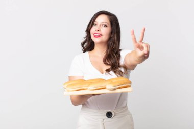 Güzel bir kadın gülümsüyor ve arkadaş canlısı görünüyor, iki numarayı gösteriyor ve elinde bir ekmek tepsisi tutuyor.